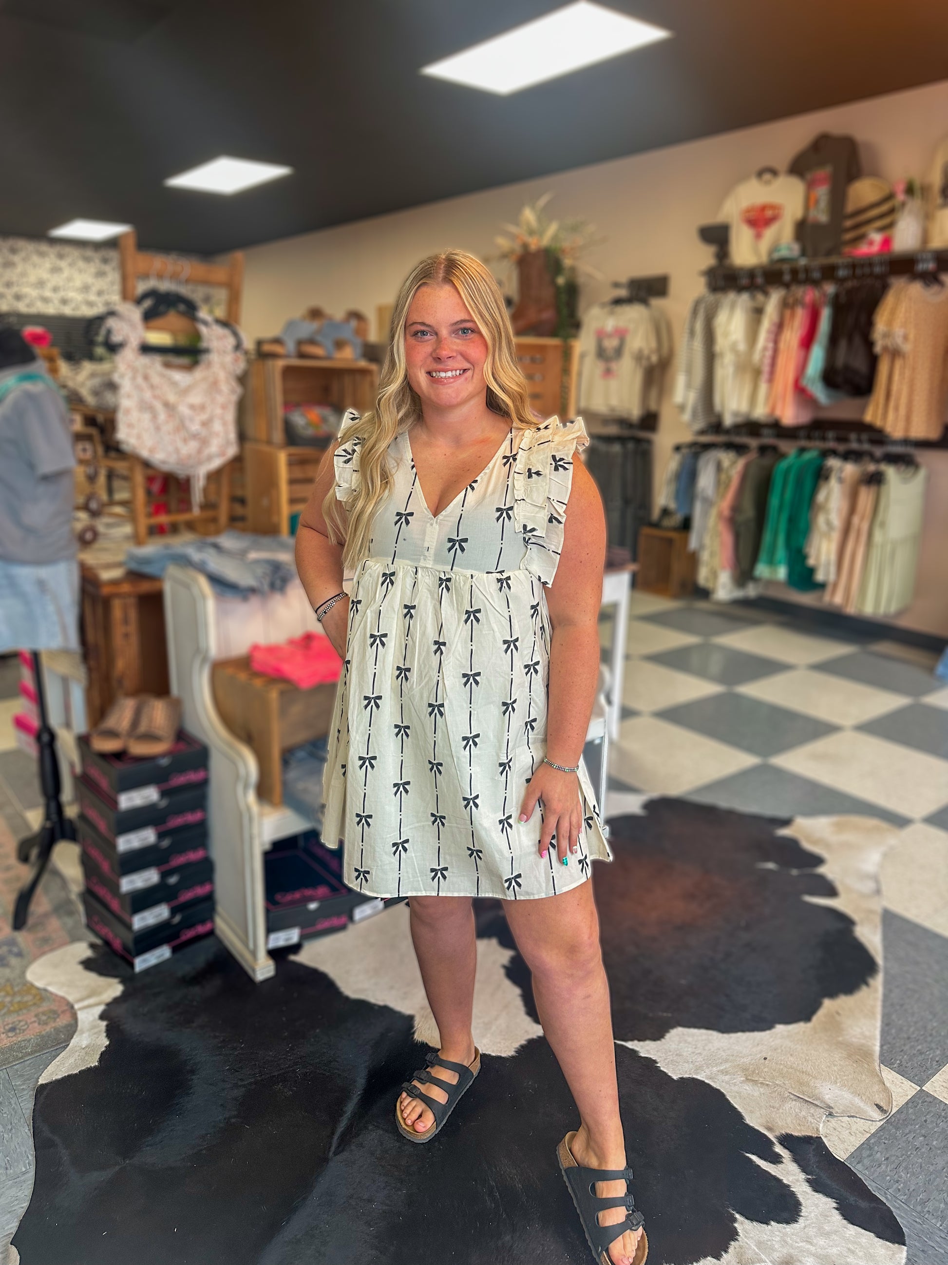 Woman wearing cream ruffle sleeve mini dress with black bow print inside a western women’s boutique with trendy clothing displays